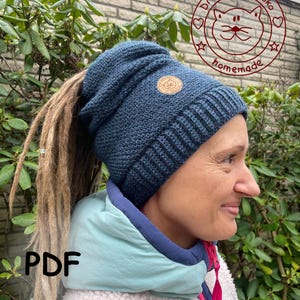 Könnte beinhalten: Eine dunkelblaue Strickmütze mit einem kleinen, runden, braunen Lederaufnäher. Die Mütze hat einen gerippten Bund und eine lockere Krone. Das Bild enthält auch den Text "PDF" und ein Logo mit dem Text "Dreos Saihoubako homemade".