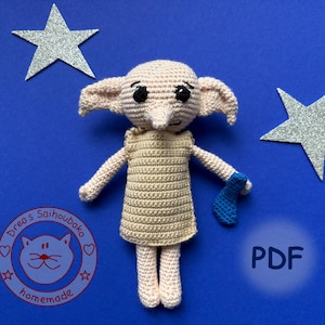 Könnte beinhalten: Eine beige Häkelpuppe mit großen Ohren, schwarzen Augen und einer spitzen Nase, die eine einfache beige Tunika trägt und eine kleine blaue Socke hält. Die Puppe ist von zwei silbernen Glitzersternen und einem runden Logo umgeben.
