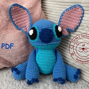Könnte beinhalten: Ein blaues, gehäkeltes Stitch-Plüschtier mit rosa Ohren und schwarzen Augen. Das Spielzeug sitzt auf einer rosa flauschigen Oberfläche und einer grauen strukturierten Oberfläche. Der Text "PDF" ist im Bild sichtbar. Der Text "Drea's Saihoubako homemade" ist in einem roten Kreis mit einem Katzenkopf darin sichtbar.