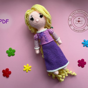 Könnte beinhalten: Eine gehäkelte Puppe von Rapunzel aus dem Disney-Film Rapunzel - Neu verföhnt. Die Puppe hat lange blonde Haare, ein lila Kleid und ein rosa Oberteil. Die Puppe liegt auf einer rosa Oberfläche mit bunten Papierblumen.