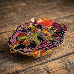 May include: A circular metal enamel pin featuring a colorful graphic of a masked character with large yellow eyes, red accents, and black details. The design includes stylized purple and orange elements surrounding the central figure with gold metal edges.