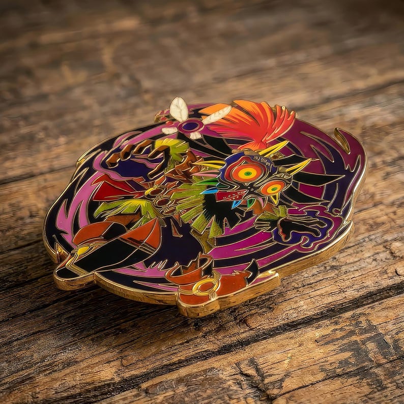 May include: A circular metal enamel pin featuring a colorful graphic of a masked character with large yellow eyes, red accents, and black details. The design includes stylized purple and orange elements surrounding the central figure with gold metal edges.