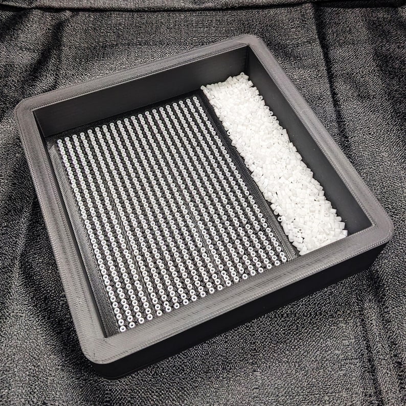 May include: A black plastic tray with a grid pattern filled with white beads. The tray is divided into two sections, one with a larger quantity of beads than the other.