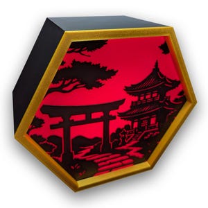 Puede incluir: Caja de luz hexagonal con marco negro y dorado, que muestra una escena de silueta japonesa. El diseño presenta una puerta torii, una pagoda y árboles, iluminados por una luz roja. Un artículo decorativo.