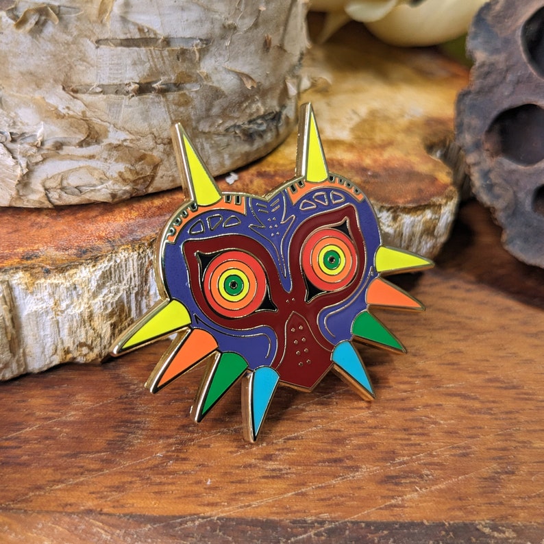 May include: A gold-colored pin with a colorful design of a mask with sharp points. The mask is red, blue, yellow, and green. The mask has two large eyes with yellow, green, and red circles.