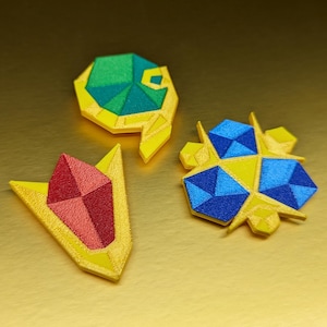 May include: Three colorful, geometric-shaped brooches. One has a red gem, another has blue and yellow shapes, and the third has green and yellow shapes. All are set against a gold background.