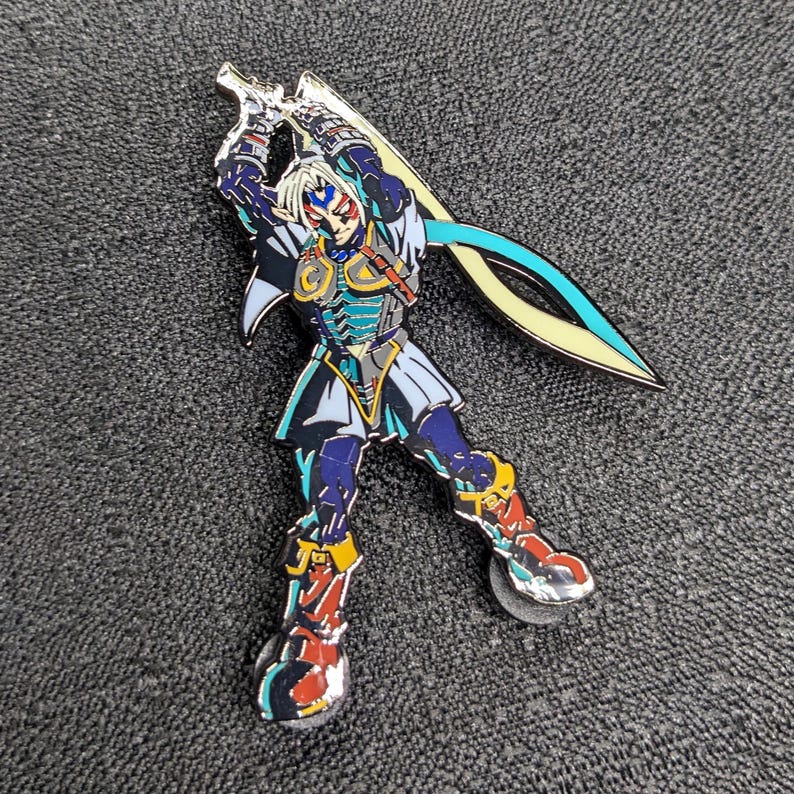 Op de afbeelding: Een blauwe en gele emaille pin met een personage uit een videogame. Het personage houdt een zwaard vast en heeft een blauwe en gele helm. De pin is een verzamelstuk voor fans van het spel.