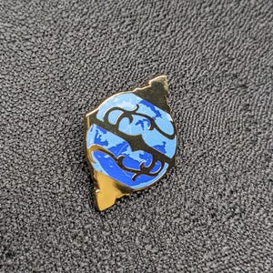 May include: A gold-colored pin with a blue and black design of a stylized globe. The globe is surrounded by a gold-colored frame.