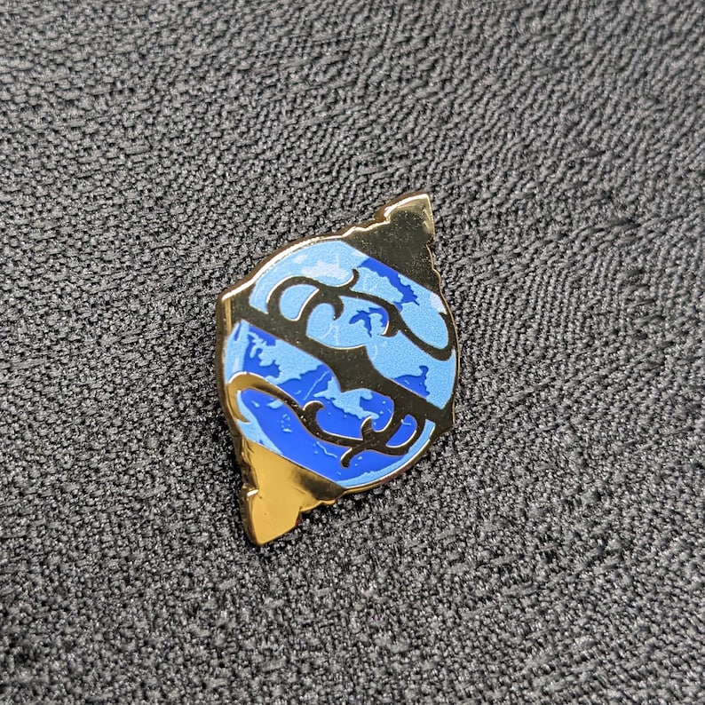 May include: A gold-colored pin with a blue and black design of a stylized globe. The globe is surrounded by a gold-colored frame.
