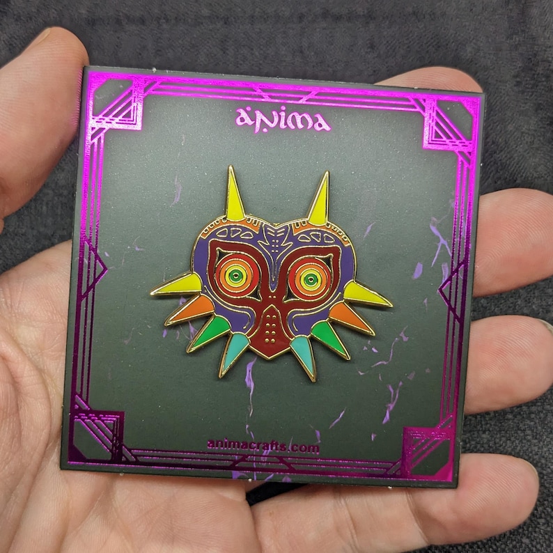 May include: A rainbow colored Majora's Mask pin on a black background with a gold border and the text "Anima" and "animacrafts.com".