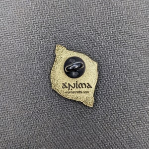 May include: The back of a gold-colored enamel pin with a black butterfly design. The pin has a black metal clasp and the text "anima animacrafts.com" is printed on the back.