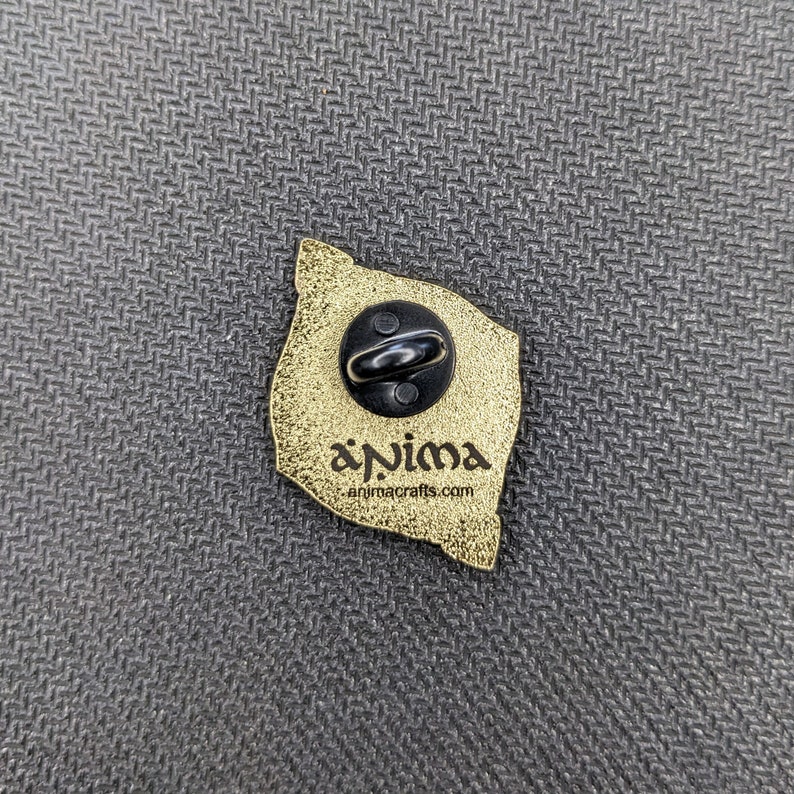 May include: The back of a gold-colored enamel pin with a black butterfly design. The pin has a black metal clasp and the text "anima animacrafts.com" is printed on the back.