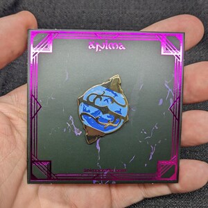 May include: A gold and blue enamel pin with a stylized design of a planet. The pin is displayed on a black background with a pink border and the text "animacrafts.com" and "anima" printed on it.