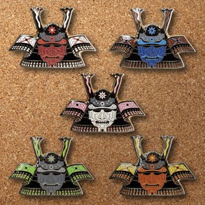 May include: Five colorful enamel pins in the shape of samurai helmets. Each pin features a different color scheme, including red, blue, white, green, and orange, with intricate details and a star emblem on the helmet.