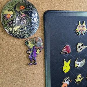 以下が含まれることがあります： ゼルダの伝説ビデオゲームシリーズのキャラクターとシンボルをフィーチャーしたエナメルピンのコレクション。ピンは、幾何学模様の黒いマウスパッドに飾られています。ピンには、緑と金のマイラのマスク、赤と白のガノンドロフ、黄色と茶色のリンク、青と白のシークが含まれています。
