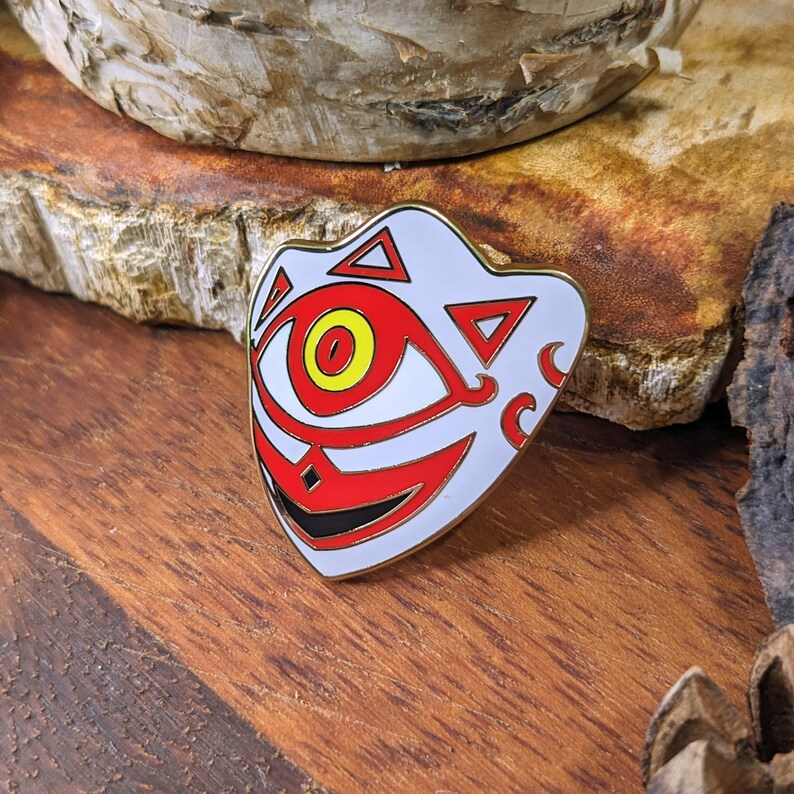 May include: White enamel pin with a red and black design. The design features a large eye with a yellow iris and a stylized face with a triangular pattern.