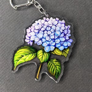 Peut inclure: Un porte-clés en acrylique transparent avec une fleur d'hortensia violette et bleue et des feuilles vertes.