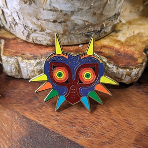 May include: A colorful enamel pin featuring the Majora's Mask from the Legend of Zelda video game series. The mask is a dark blue with red and yellow accents, and has a sharp, pointed design.