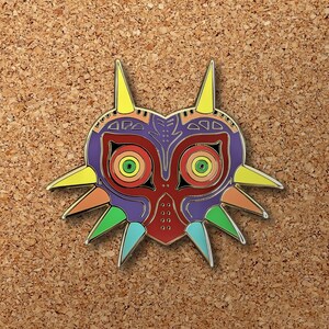 May include: A colorful enamel pin featuring the Majora's Mask from the Legend of Zelda video game series. The mask is purple, red, and green with yellow and orange accents.