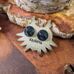 May include: Gold-colored metal pin with a stylized animal face design. The pin has two black metal clasps on the back and the text "a'Nima" is printed on the back.
