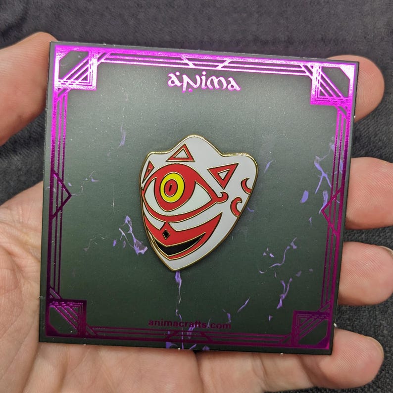 May include: A white enamel pin with a red and yellow design of a mask with a single eye. The pin is on a black background with a pink border and the text "anima" and "animacrafts.com".