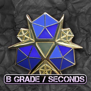 Può includere: Spilla geometrica con contorno color oro e forme triangolari blu e verdi. La spilla ha un design a forma di stella con il testo "B GRADE / SECONDS" in bianco su sfondo nero.