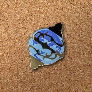 May include: A blue and black enamel pin with a stylized design of a wave or ocean. The pin has a shiny, metallic finish.