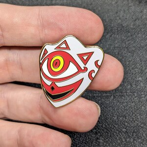 May include: A white enamel pin with a red and yellow eye, black and red mouth, and gold outline. The pin is shaped like a mask with a geometric design.