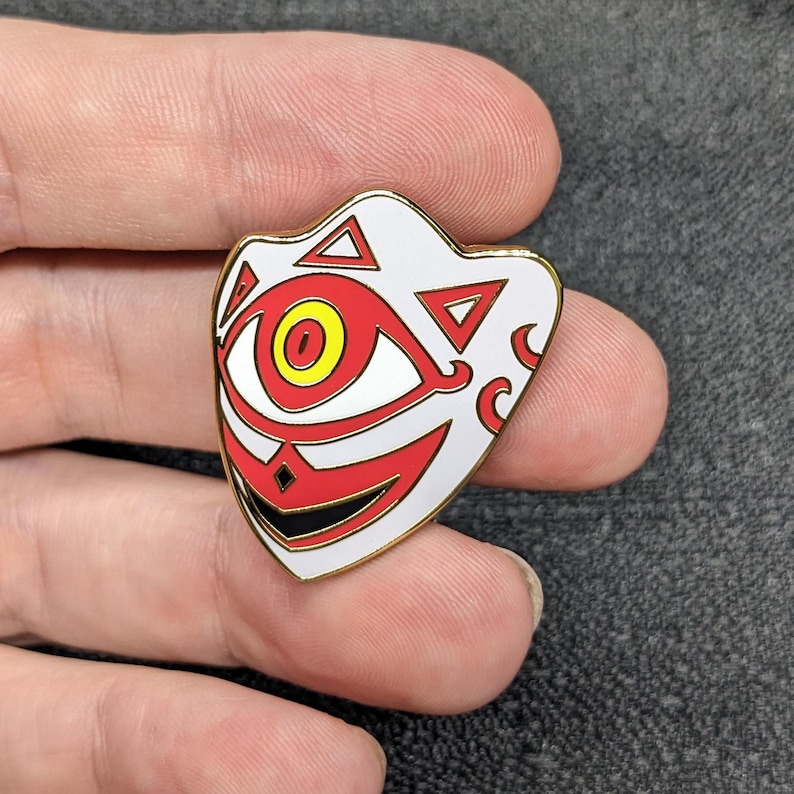 May include: A white enamel pin with a red and yellow eye, black and red mouth, and gold outline. The pin is shaped like a mask with a geometric design.