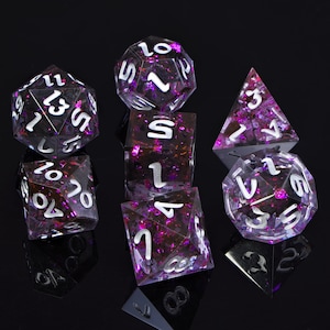 May include: A set of seven purple and silver dice with iridescent glitter. The dice are arranged in a pyramid shape on a black surface.