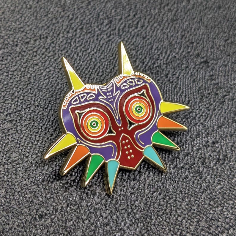 May include: A colorful enamel pin featuring the Majora's Mask from the Legend of Zelda video game series. The mask is a stylized, cartoonish depiction of a demonic face with sharp teeth and horns. The mask is painted in a variety of colors, including red, orange, yellow, green, blue, and purple.