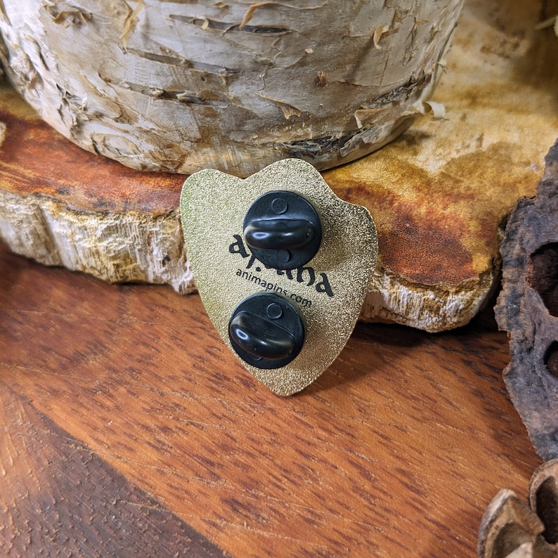 May include: Gold-colored enamel pin with a black and white illustration of a wolf's head. The pin has two black rubber backs. The text "animapins.com" is printed on the pin.
