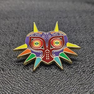 May include: A gold-colored enamel pin featuring a colorful Majora's Mask from the Legend of Zelda video game series. The mask has a purple, red, and green design with yellow accents.