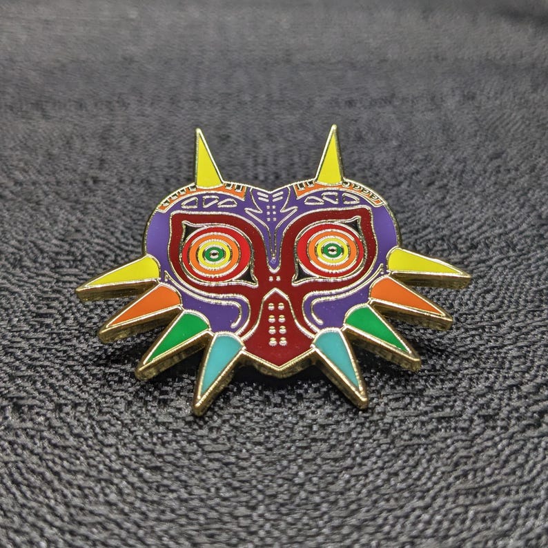 May include: A gold-colored enamel pin featuring a colorful Majora's Mask from the Legend of Zelda video game series. The mask has a purple, red, and green design with yellow accents.