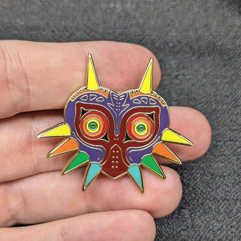 May include: A colorful enamel pin featuring the Majora's Mask from the Legend of Zelda video game series. The mask is red, purple, green, yellow, and orange with gold accents.
