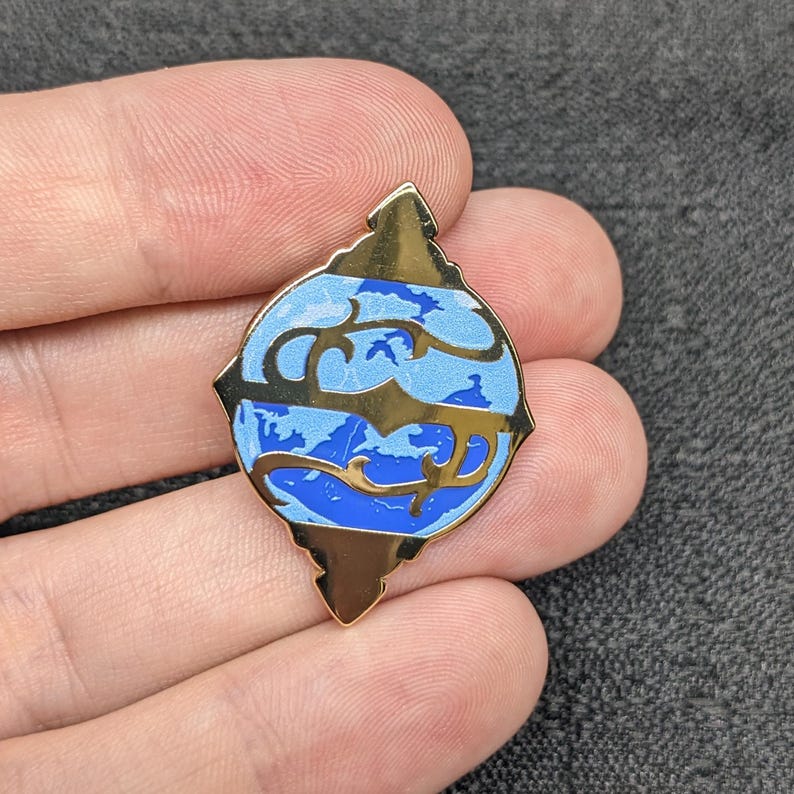 May include: A gold-colored pin with a blue and white enamel design. The design features a stylized globe with a swirling pattern.