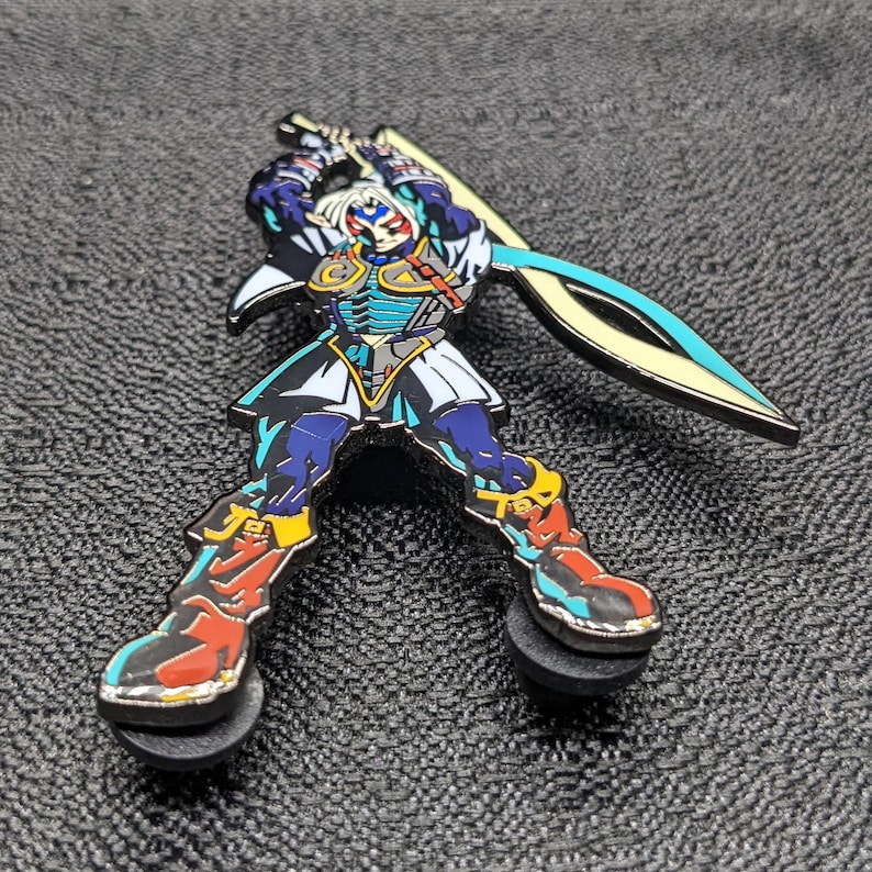 Op de afbeelding: Een blauwe en gouden emaille pin van een personage uit een videogame. Het personage houdt een zwaard vast en heeft een blauw en wit masker. De pin is op een zwarte achtergrond.