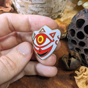 May include: A white enamel pin with a gold outline, depicting a stylized eye with red and black details. The eye is surrounded by a white mask with a red and black design.