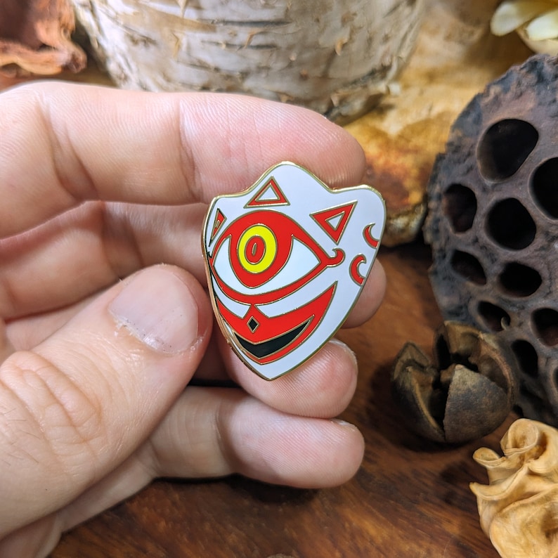 May include: A white enamel pin with a gold outline, depicting a stylized eye with red and black details. The eye is surrounded by a white mask with a red and black design.