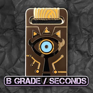 May include: A brown and gold metal pin with a stylized eye design and a blue iris. The rectangular pin has a handle at the top and gold accents. The text "B GRADE / SECONDS" is displayed at the bottom of the pin.