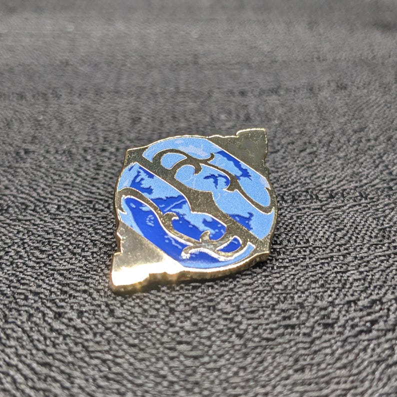 May include: A gold-colored pin with a blue and white design. The design features a stylized wave pattern with a white outline.