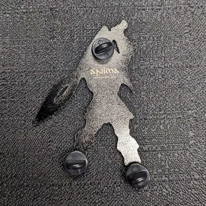 Op de afbeelding: Een zilverkleurige metalen pin met een gestileerd figuur dat een zwaard vasthoudt. De pin heeft een zwarte achterkant met drie zwarte pin-ruggen. De pin heeft de tekst "Anima" in gouden letters.