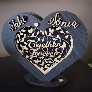 May include: A wooden heart-shaped sign with the names "Jake" and "Sonia" on either side. The center of the heart features a cutout design of leaves and vines with the words "Together Forever" in a decorative font.