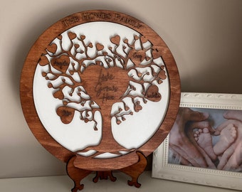 May include: A wooden family tree plaque with the text "The Howes Family" at the top. The tree has branches with hearts that have names written on them, including "Angie", "Cory", "Lila", and "Alice". The tree is on a wooden stand.
