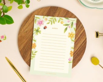 May include: A floral-themed notepad with the word "notes" at the top. The notepad features a border of pink, orange, and yellow flowers, green leaves, and a ladybug. The notepad is on a wooden cutting board.