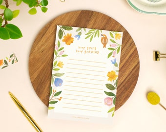 May include: A floral-themed notepad with the words "keep going keep growing" in gold script. The notepad is surrounded by watercolor flowers in shades of yellow, blue, and pink. The notepad is on a wooden surface.