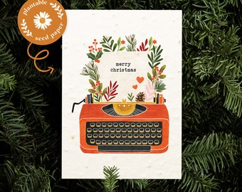 May include: A Christmas card featuring a vintage orange typewriter illustration with the words "merry christmas". The typewriter is decorated with a wreath of leaves, berries, and hearts. The card is made of plantable seed paper.
