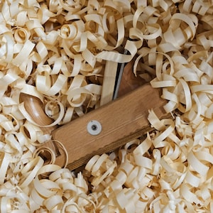 Könnte beinhalten: Ein hölzerner Hobel mit einer Metallklinge ist von Holzspänen umgeben. Der Hobel ist aus hellem braunem Holz gefertigt und hat einen Metallknopf an der Seite.