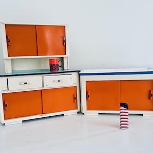 Könnte beinhalten: Ein Satz Vintage-Küchenschränke mit orangefarbenen Schiebetüren und weißen Rahmen. Das Set umfasst einen Wandschrank, einen Unterschrank mit Schubladen und einen separaten Schrank. Ein roter Becher und ein gestreiftes Feuerzeug sind ebenfalls vorhanden.