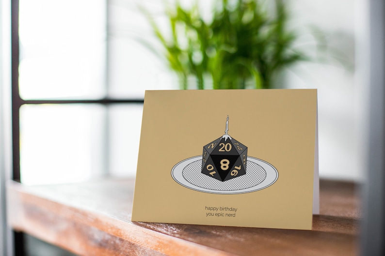 Pu&ograve; includere: Un biglietto beige con un dado nero e oro a 20 facce con una candela accesa in cima. Il dado &egrave; su un piatto bianco con un motivo bianco e nero. Il testo sul biglietto recita "happy birthday you epic nerd".